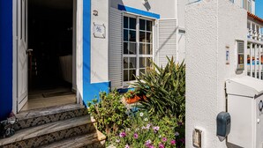 A building with a white exterior and blue trim, a door leading to an interior, and a small garden with various plants. - 'Alojamento Local Família Coelho' House with Wi-Fi (Quinta do Conde)