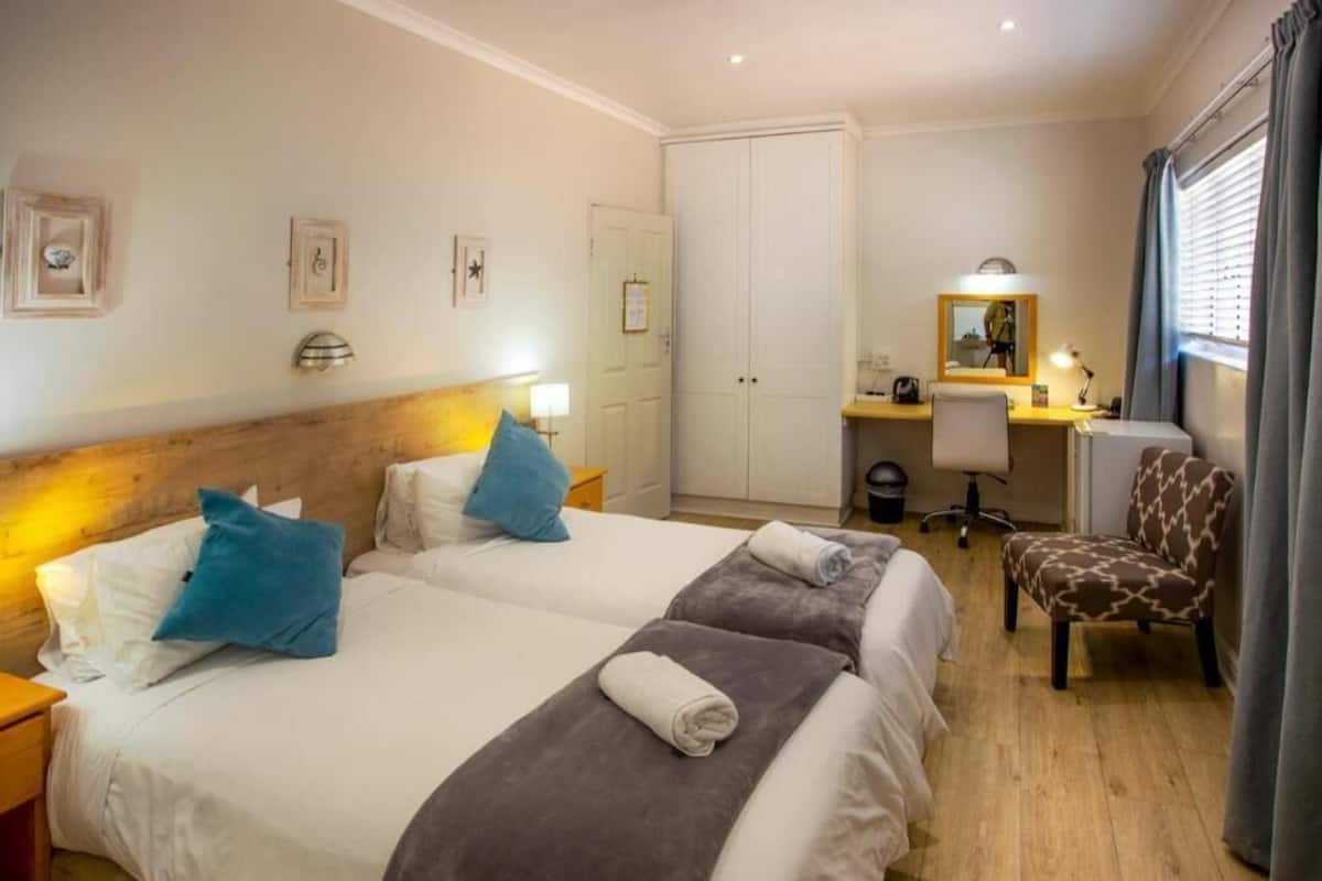 Quarto de hotel com duas camas, uma escrivaninha, uma cadeira, um guarda-roupa e um espelho.