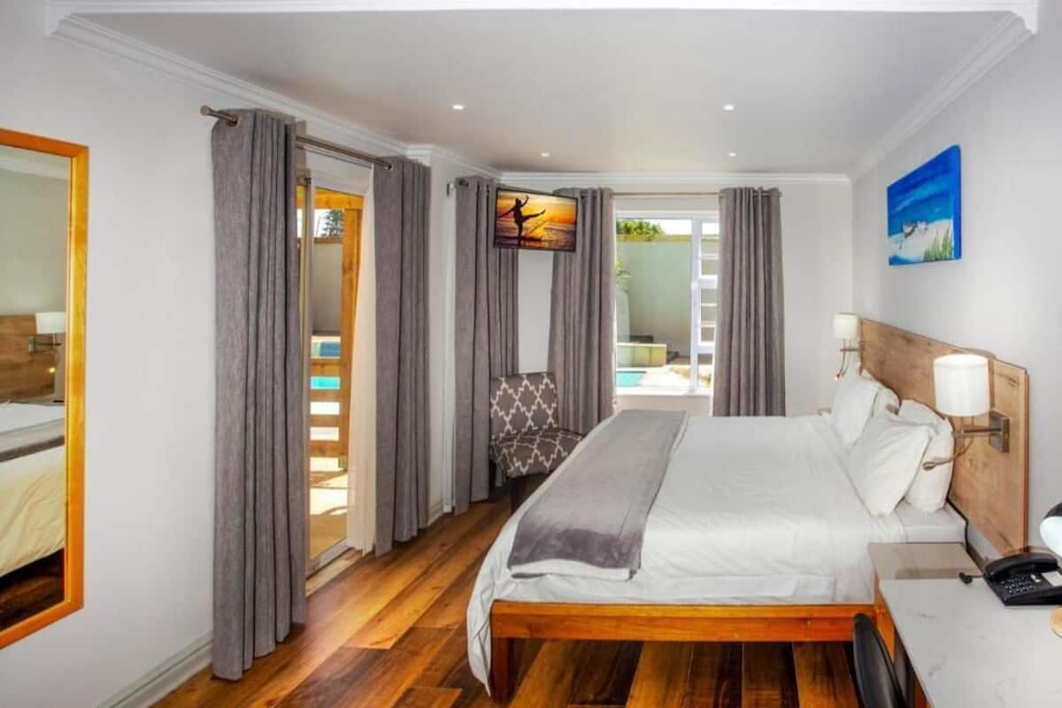 Quarto de hotel com cama de madeira, uma escrivaninha, uma cadeira, mesa de cabeceira, espelho e vista para a piscina.