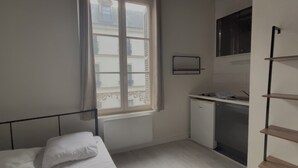 A small, compact room with a bed, a desk with a computer, a small refrigerator, and a window with a view of buildings. - Beautiful Studio T1 in Vieux tours (tours)