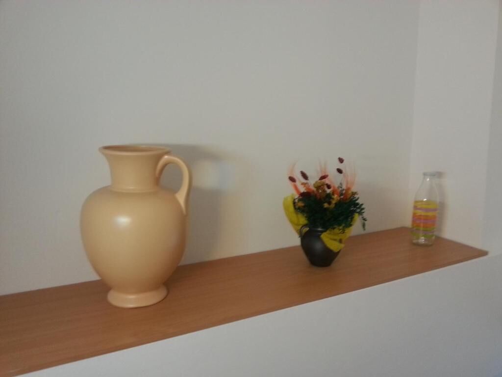 Un jarrón color beige, una planta pequeña en maceta y una botella a rayas sobre una estantería de madera.