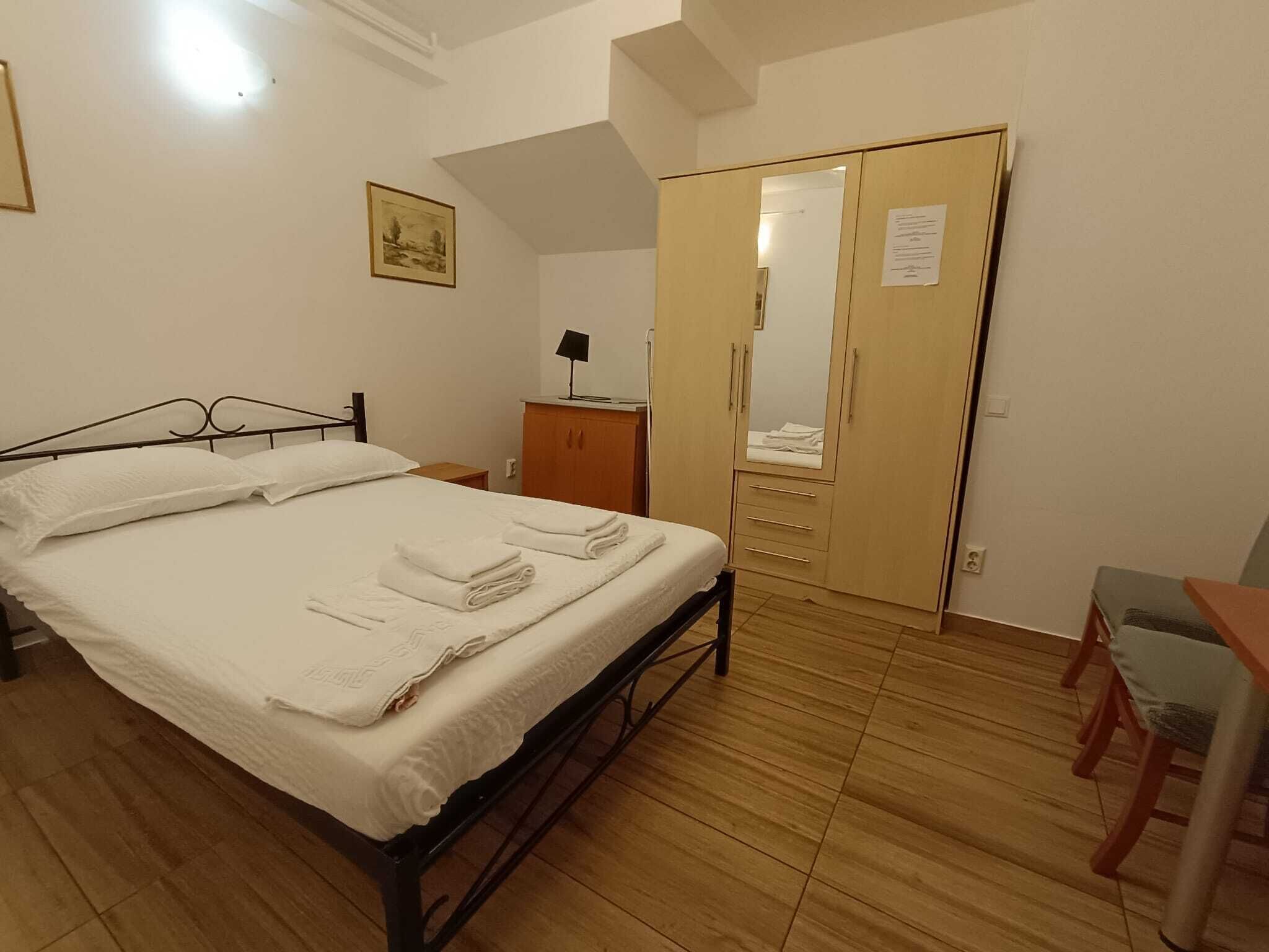 Ein ordentlich bezogenes Bett mit gefalteten Handtüchern, ein Holzschrank mit Spiegel, ein kleiner Tisch mit Lampe und ein Stuhl.