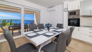Una cocina moderna con horno empotrado y microondas, un comedor con mesa dispuesta para cuatro personas, y un amplio ventanal corredero que da a un balcón con vistas al mar.