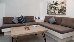 A living room with a brown sectional sofa, a wooden coffee table, and a tropical plant wall art. - mati cave villas (Santorini)