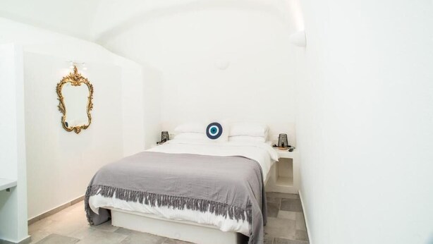 A bedroom with a bed, bedside tables, a wall-mounted mirror, and a decorative wall piece. - mati cave villas (Santorini)