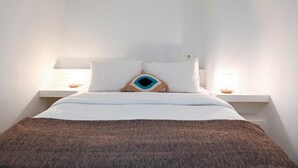 A bedroom with a bed, pillows, a blanket, bedside tables, and a wall-mounted light fixture. - mati cave villas (Santorini)