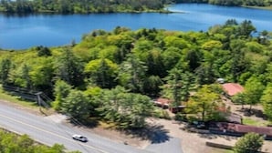 En sjö omgiven av tät skog, en väg med ett fordon på och en liten byggnad med ett rött tak.