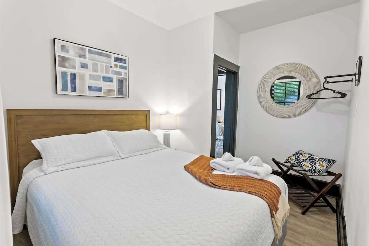 Una cama bien hecha con cabecera, un espejo montado en la pared, un perchero para toallas y una obra de arte abstracta enmarcada.