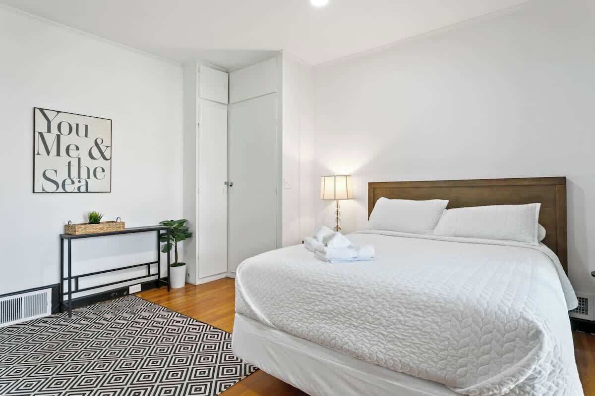 Une chambre à coucher bien rangée, avec un lit, une table de chevet, une lampe et un mur orné d’une citation encadrée.