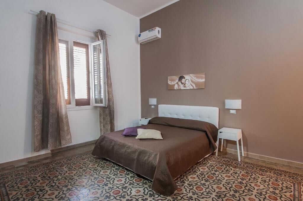 Un dormitorio con cama, suelo estampado, ventana con cortinas, aire acondicionado de pared y un cuadro.