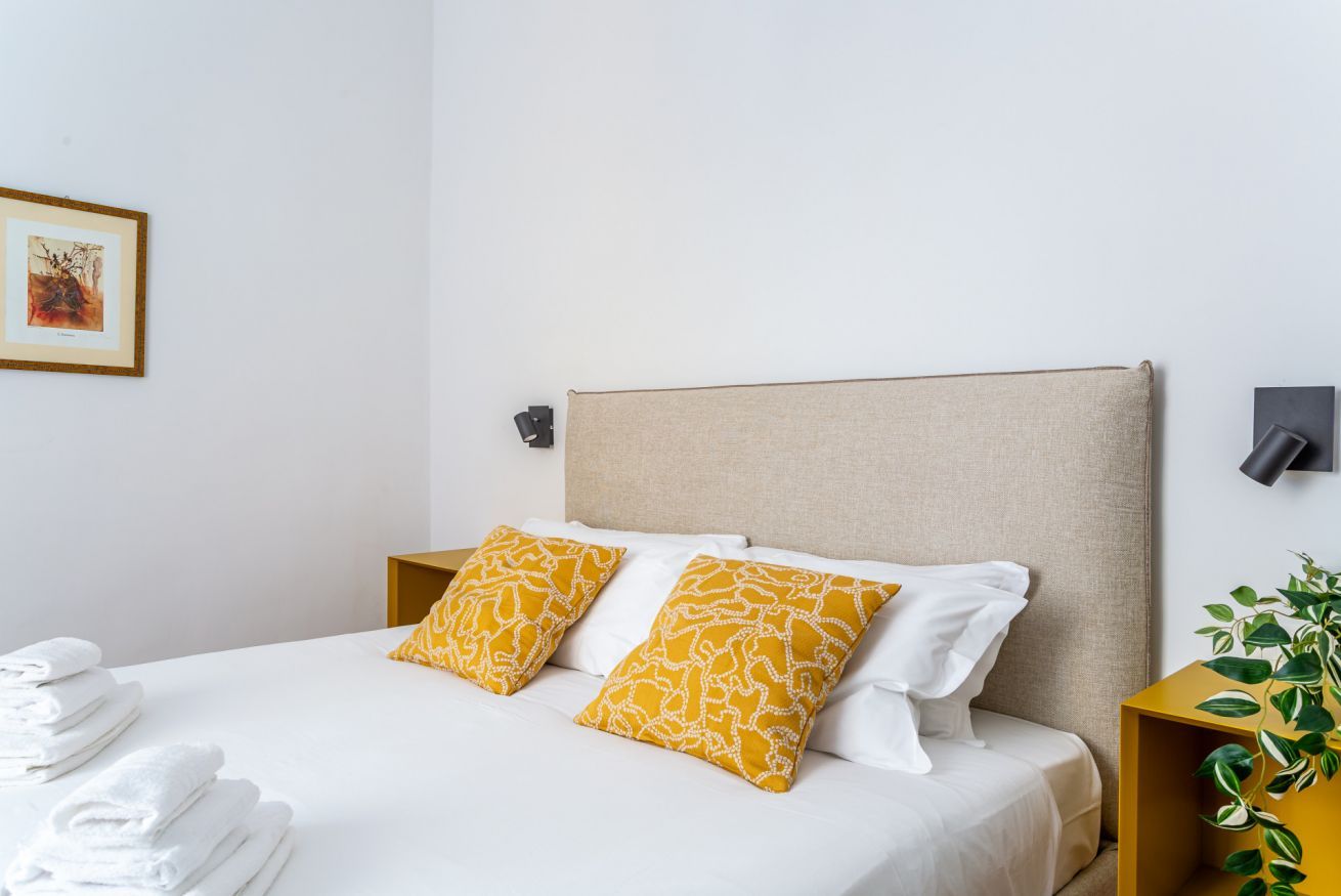 A neatly made bed with white linens and two patterned pillows, a small bedside table with a lamp, and a framed picture on the wall.