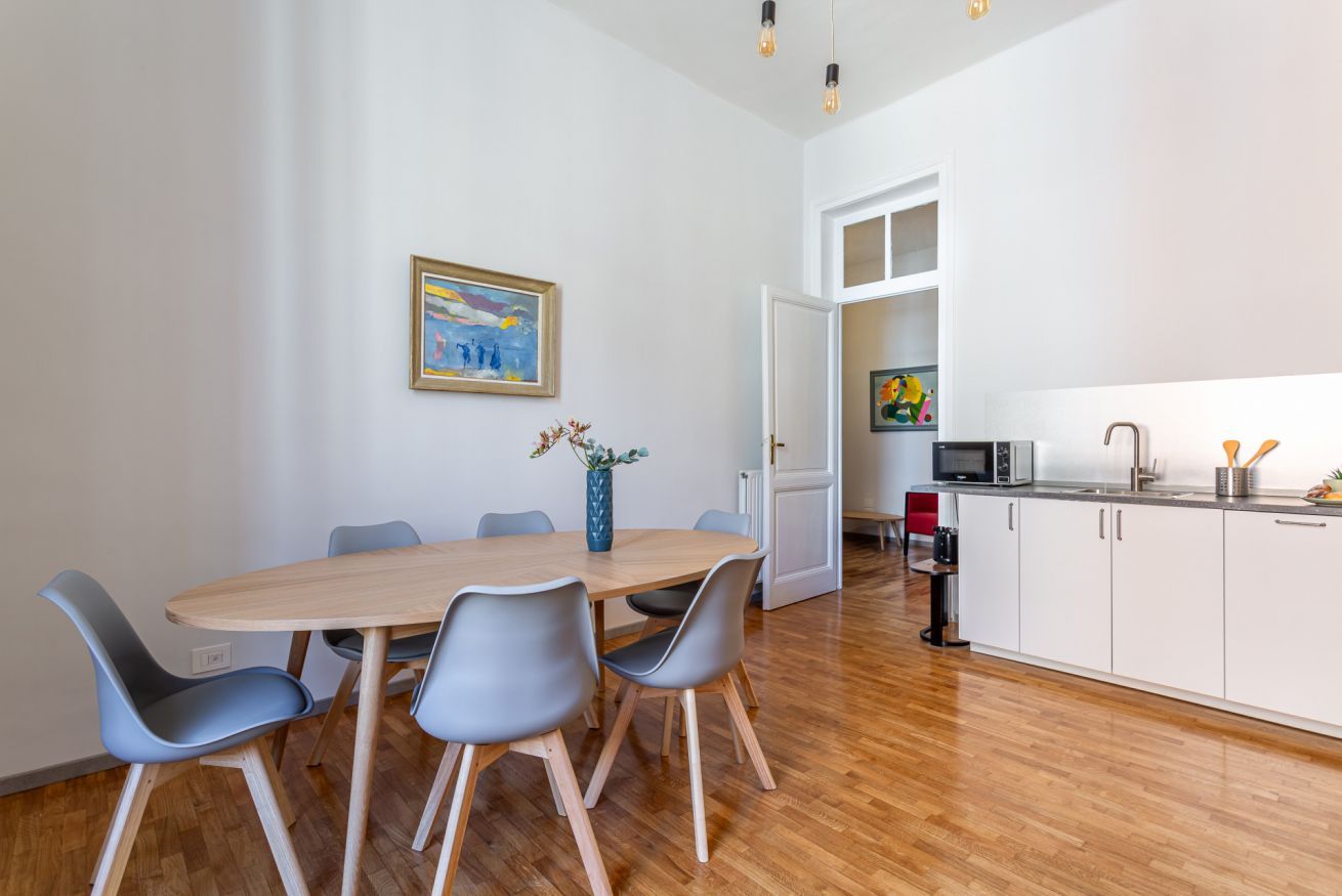 A dining area with a wooden table and grey chairs, a painting on the wall, and a kitchen in the background.
