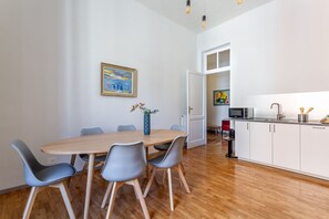 Un comedor con mesa de madera y sillas grises, un cuadro en la pared y una cocina al fondo.