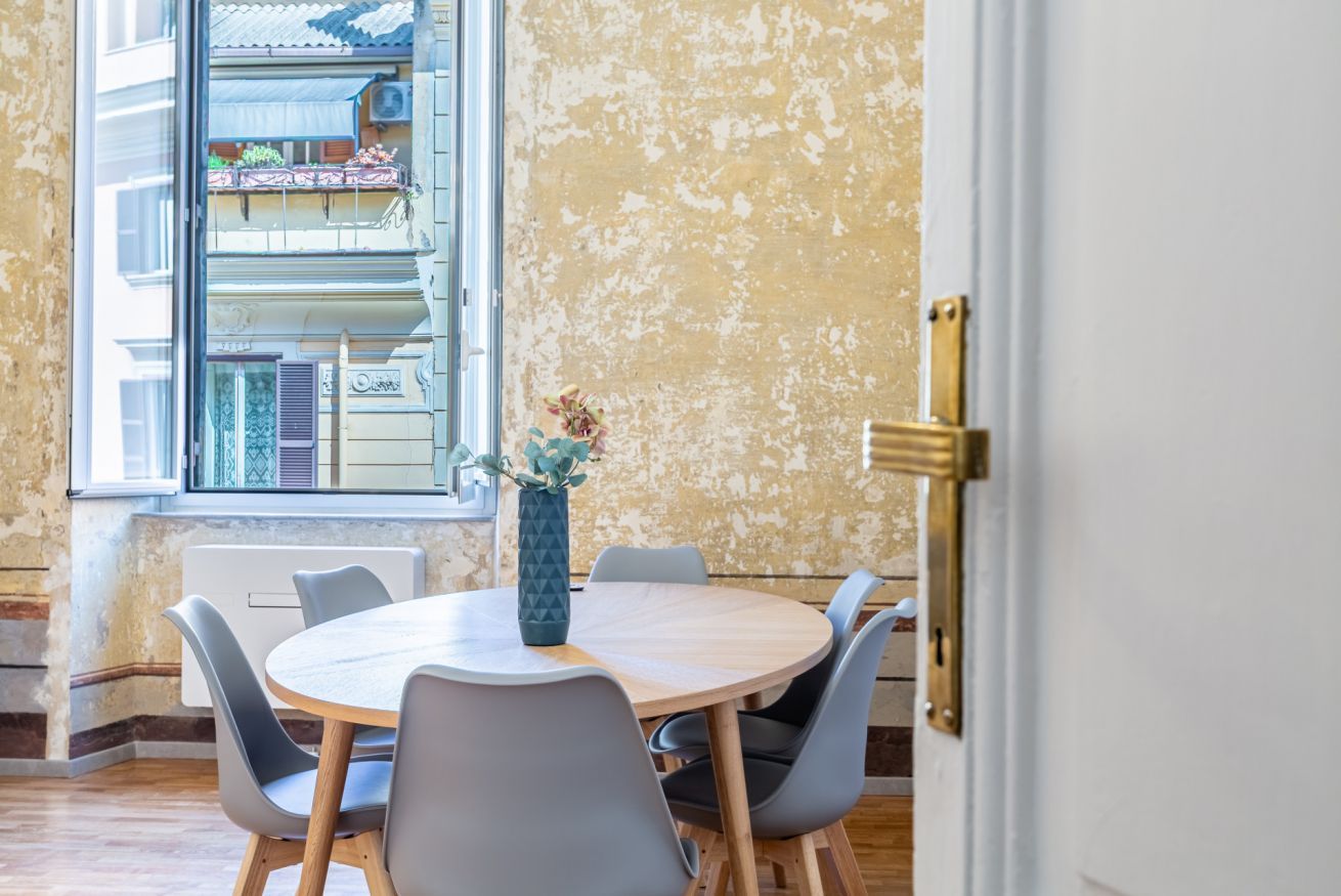 A round dining table with chairs, a vase with flowers, and a window with a view of a building.