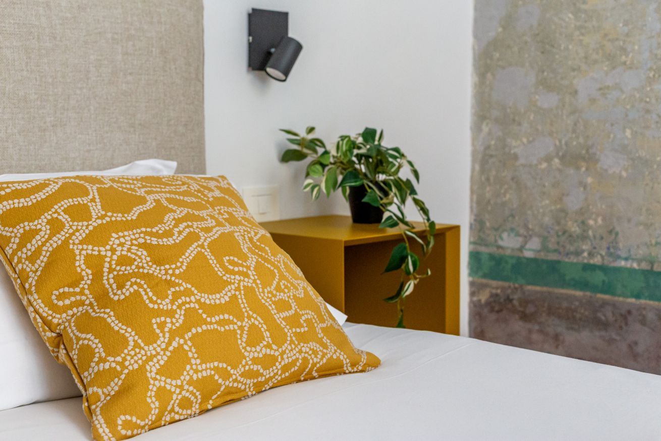 A bed with a white pillow and a mustard-colored pillow with a white pattern, a small wooden nightstand with a plant, and a wall-mounted light fixture.