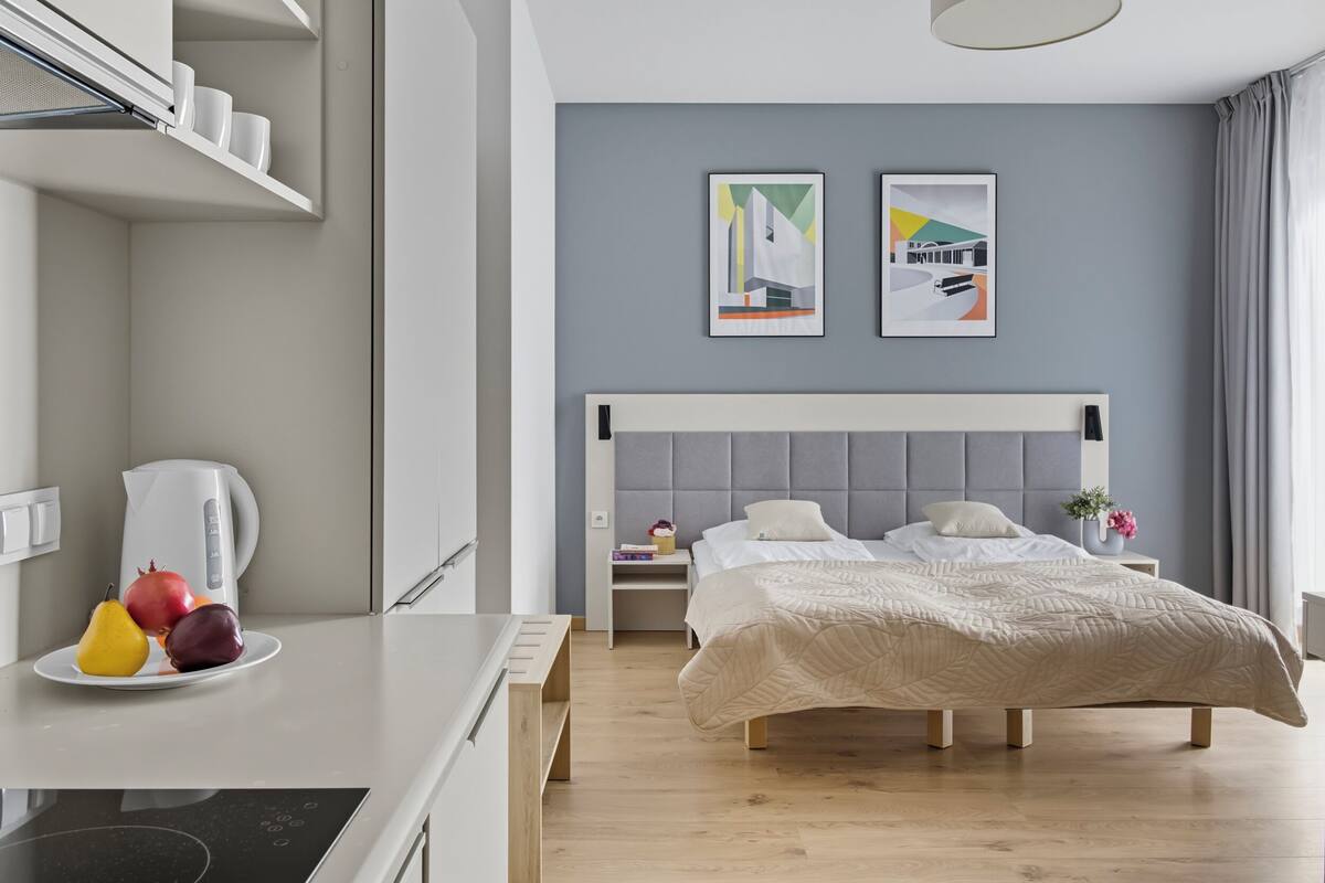 A modern hotel room with a bed, bedside tables, a desk with a kettle and fruit, and artwork on the wall.