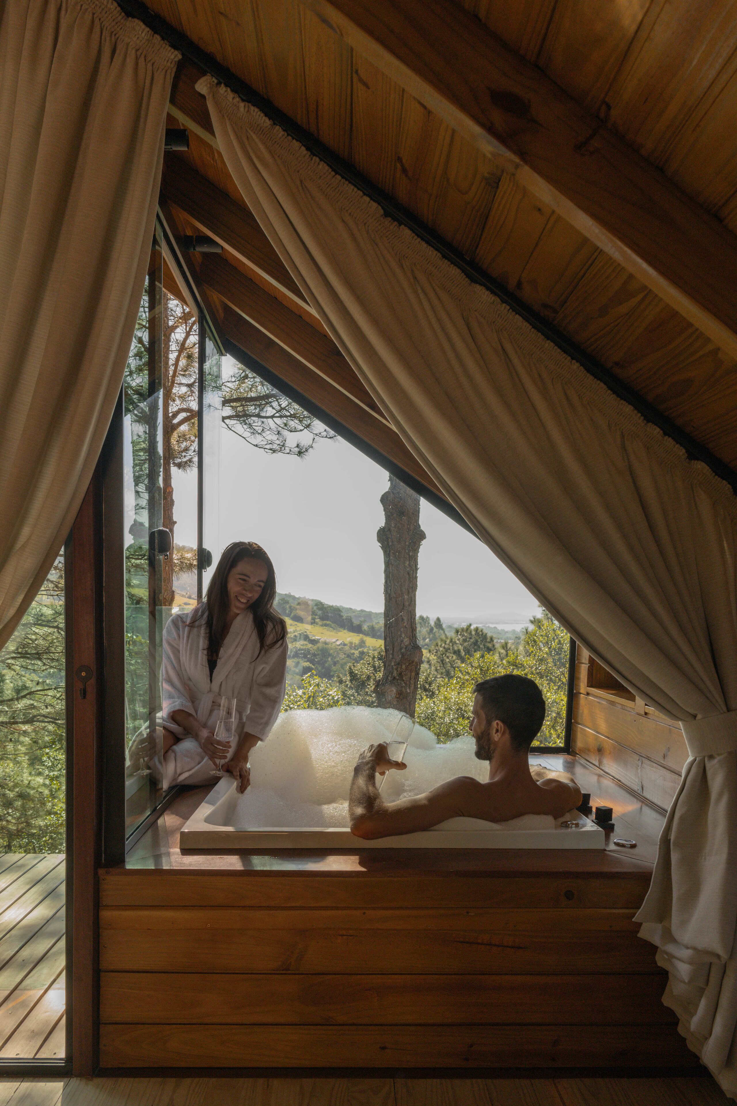A wooden cabin with a glass-enclosed hot tub, a wooden deck, and a view of the surrounding forest.