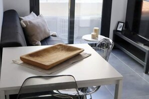Una sala moderna con una mesa blanca, un sofá gris, un ventanal que da al exterior y un televisor a la derecha.