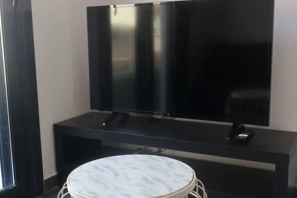 A modern living room with a flat-screen TV on a black stand, a round white ottoman, and a dark-colored sofa.