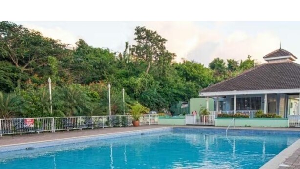 A swimming pool with a tiled deck, surrounded by greenery and a building with a tiled roof. - COMFORTABLE KING BEDROOM FLAT in townhome in a pleasant Hilltop neighborhood, (Kingston 6)