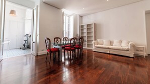 Un vaste espace de vie doté d’un plancher en bois, d’une table à manger avec un plateau en verre et quatre chaises, d’un canapé blanc et d’une bibliothèque.
