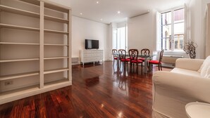 Un vaste espace de vie doté d’un plancher en bois, d’un canapé blanc, d’une table à manger avec des chaises et d’un téléviseur posé sur un meuble blanc.