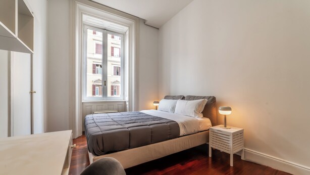 Une chambre à coucher avec un lit, une table de chevet, une lampe et une vue sur les bâtiments par la fenêtre.