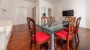 Un coin repas avec une table en verre et des chaises en bois, un canapé blanc et un téléviseur fixé au mur.