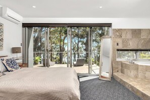 Ein modernes Schlafzimmer mit einem großen Bett, ein Badezimmer mit einer Badewanne und eine Schiebeglastür zu einem Außenbereich mit Blick auf Bäume.