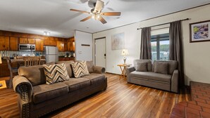 A living room with a brown sofa, a grey sofa, a wooden floor, a ceiling fan, a kitchen with wooden cabinets, and a window with curtains. - Whispering Pines | 3 Bedroom Suite | Pet Friendly! (Sevierville)