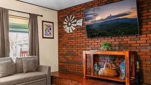A living room with a brick wall, a sofa, a TV mounted on the wall, a wooden console table with decorative items, and a window with curtains. - Whispering Pines | 3 Bedroom Suite | Pet Friendly! (Sevierville)