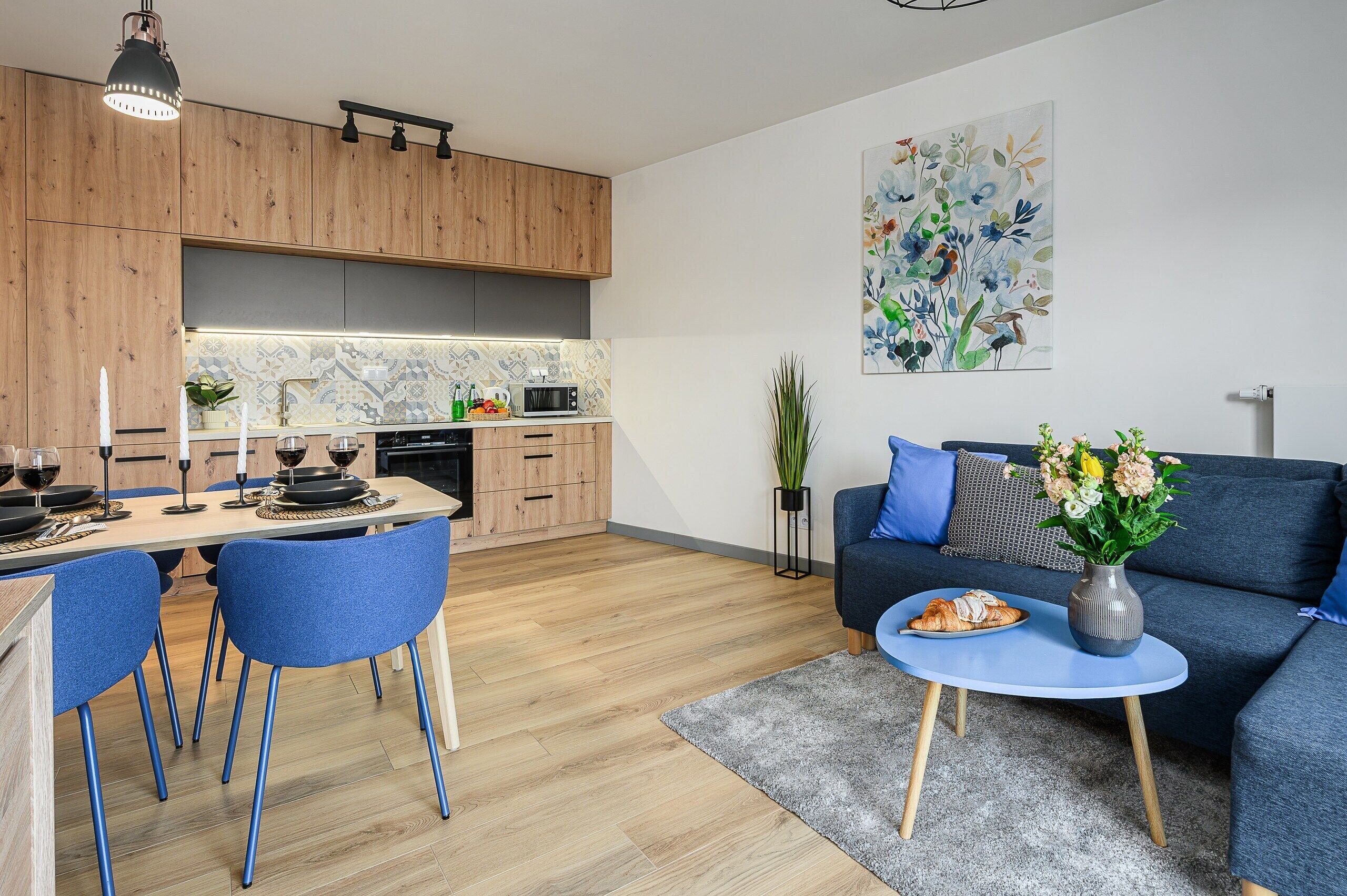 Una cocina moderna con armarios de madera, un área de comedor con sillas azules, un sofá y una mesita con un jarrón.