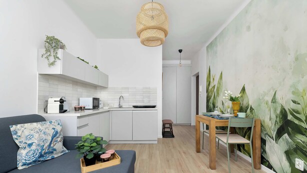 Et moderne køkken med hvide skabe, et spiseområde med et træbord og stole, samt en opholdsstue med sofa og pyntepuder.