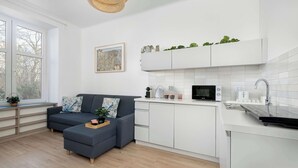Et moderne køkken med hvide skabe, en mikrobølgeovn og en vask. Der er en mørkegrå sofa med en plante og et billede i ramme på væggen.