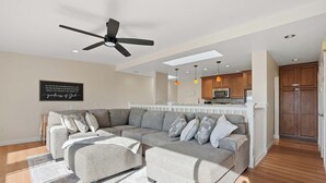 Un salon moderne avec un canapé d’angle, un ventilateur de plafond, un éclairage encastré et une cuisine en arrière-plan.