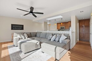Ein modernes Wohnzimmer mit einem Ecksofa, Deckenventilator, Einbauleuchten und einer Küche im Hintergrund.