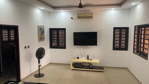 Una sala de estar con un televisor de pantalla plana, un ventilador de techo, un ventilador de pie, un mueble para televisión y una ventana con diseño de cuadrícula.
