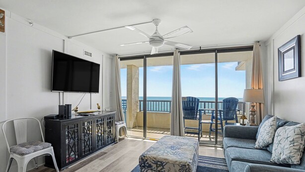 A modern living room with a flat-screen TV, a ceiling fan, a sofa, a coffee table, and a balcony with a view of the ocean. - Sea Winds #502-Oceanfront Oasis with Indoor Pool! In Windy Hill - NMB! (North Myrtle Beach)