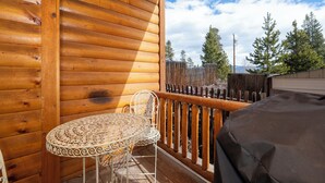 A wooden deck with a table and chairs, a covered grill, and a view of trees and mountains.