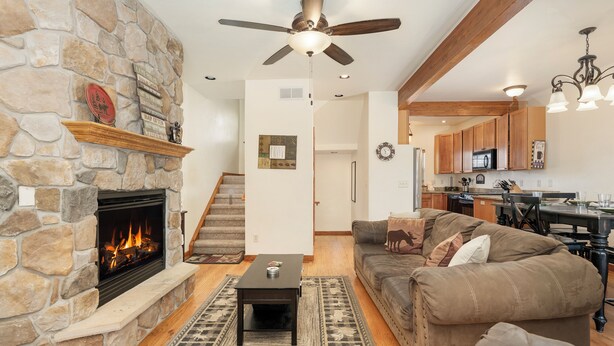 A living room with a stone fireplace, a ceiling fan, a sofa, and a kitchen in the background.