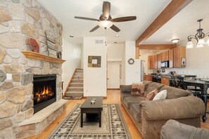 A living room with a stone fireplace, a ceiling fan, a sofa, and a kitchen in the background.