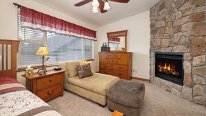 A bedroom with a stone fireplace, a ceiling fan, a wooden dresser, a sofa, and a bedside table with a lamp.