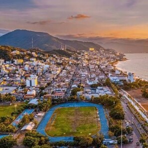 Une vue aérienne d’une ville côtière, avec un grand stade, des immeubles résidentiels et une montagne en arrière-plan.