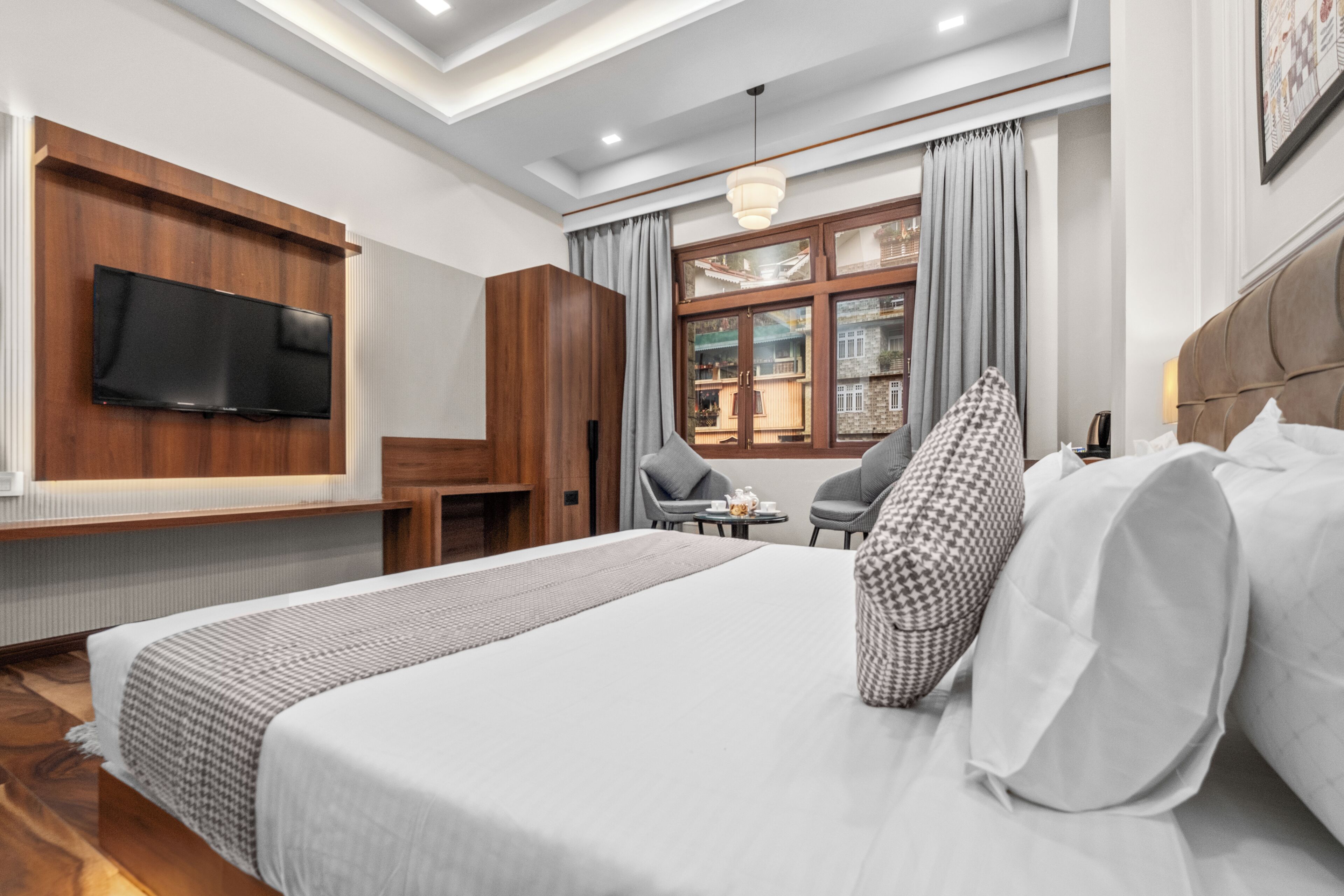 A hotel room with a bed, a TV mounted on a wooden panel, a window with curtains, and a small table with chairs.