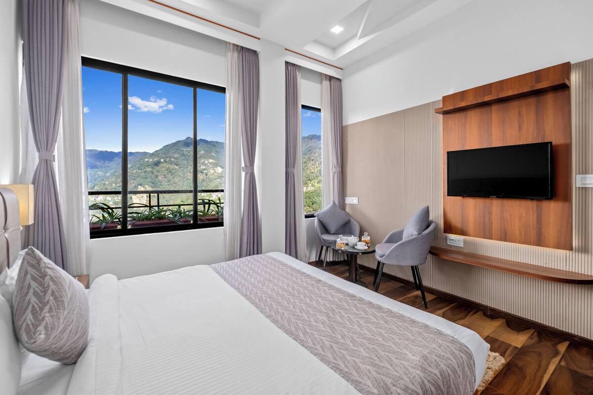 Een hotelkamer met een groot bed, een aan de muur bevestigde televisie, een klein tafeltje met stoelen en uitzicht op bergen door grote ramen.