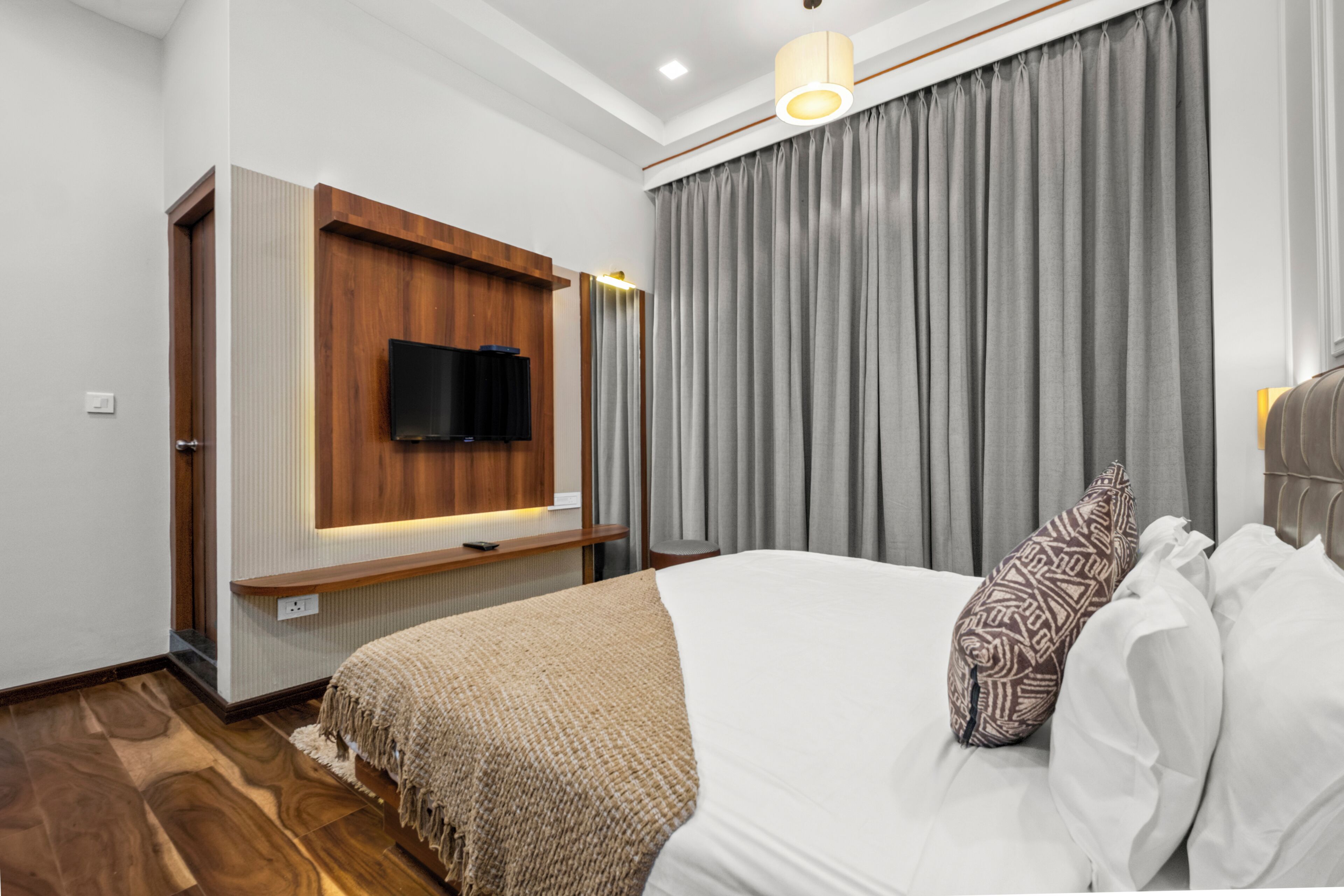 A modern hotel room with a flat-screen TV mounted on a wooden panel, a large bed with white linens, and a grey curtain.