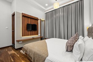 A modern hotel room with a flat-screen TV mounted on a wooden panel, a large bed with white linens, and a grey curtain.