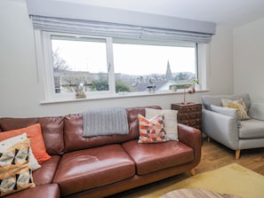 Sala de estar com um sofá de couro marrom, uma poltrona cinza e vista para o horizonte urbano através de uma grande janela.