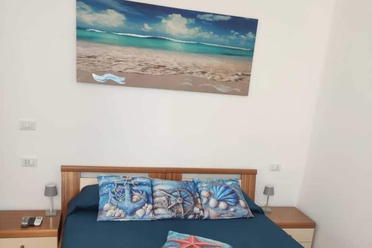 A bedroom with a bed, a nightstand, a lamp, and a painting of a beach scene.