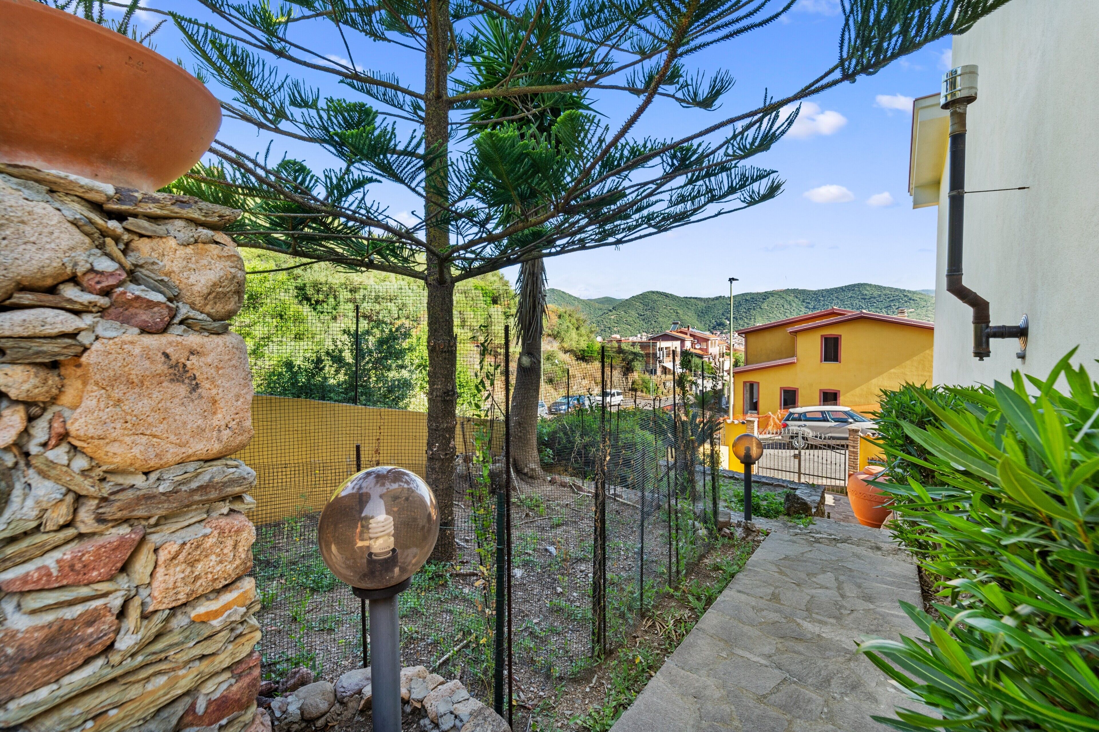 A stone wall, a metal fence, a tree, a yellow building, and a clear sky.